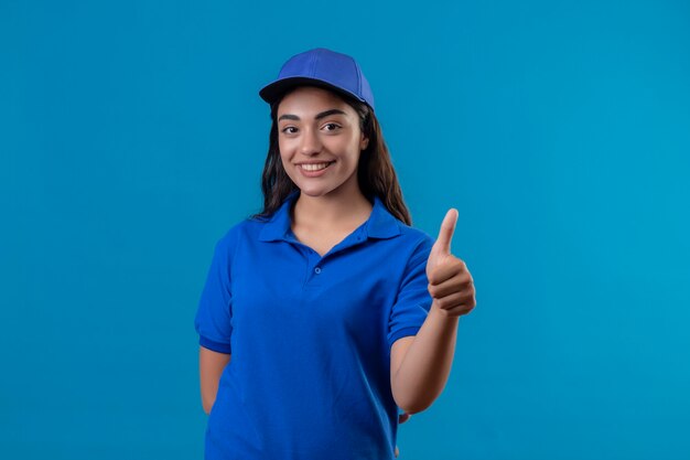 Young delivery girl in blue uniform and cap looking at camera smiling friendly happy and positive showing thumbs up standing over blue background