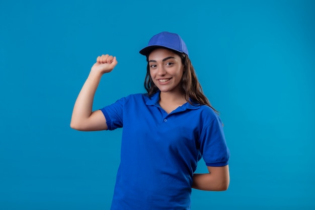 Young delivery girl in blue uniform and cap raising fist looking at camera smiling confident rejoicing her success and victory standing over blue background