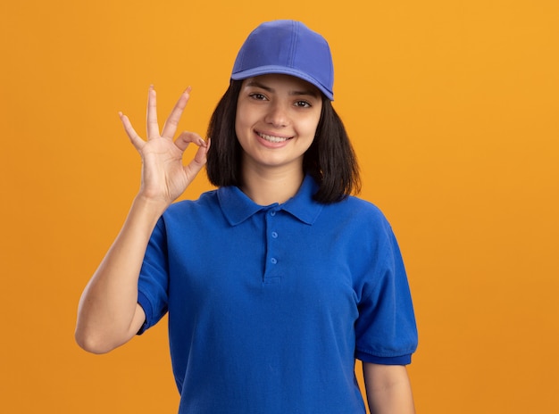 Young delivery girl in blue uniform and cap  sniling showing ok sign standing over orange wall