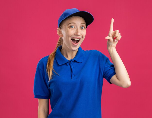 Free Photo young delivery girl in blue uniform and cap  surprised and happy showing index finger having new idea standing over pink wall