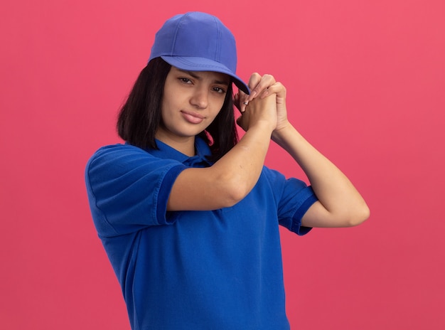 Young delivery girl in blue uniform and cap  with serious face making team gesture standing over pink wall