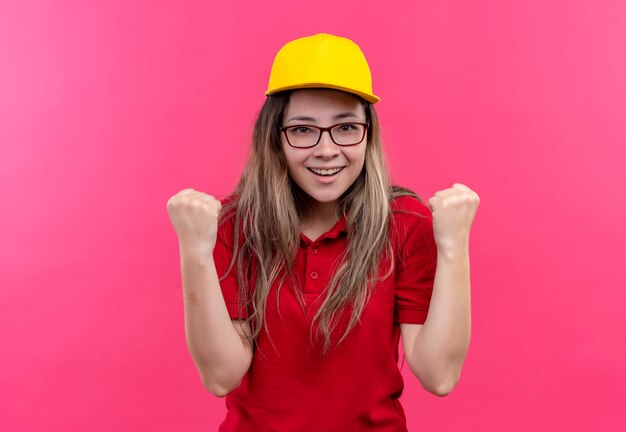 Young delivery girl in red polo shirt and yellow cap exited and happy clenching fists 