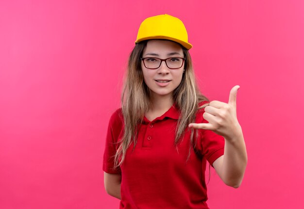 Young delivery girl in red polo shirt and yellow cap smiling making call me gesture 