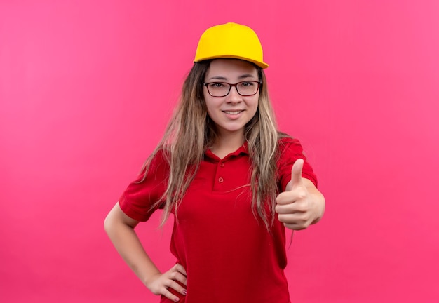 Young delivery girl in red polo shirt and yellow cap smiling showing thumbs up 