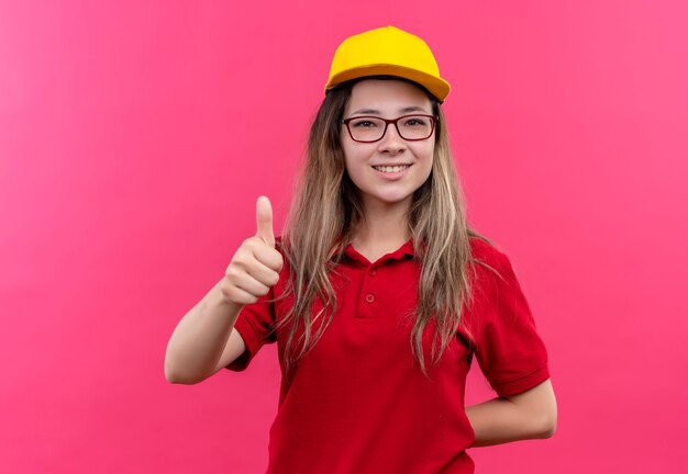 Young delivery girl in red polo shirt and yellow cap smiling showing thumbs up 