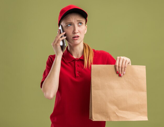 Young delivery girl in red uniform and cap holding paper package looking confused while talking on mobile phone over green wall