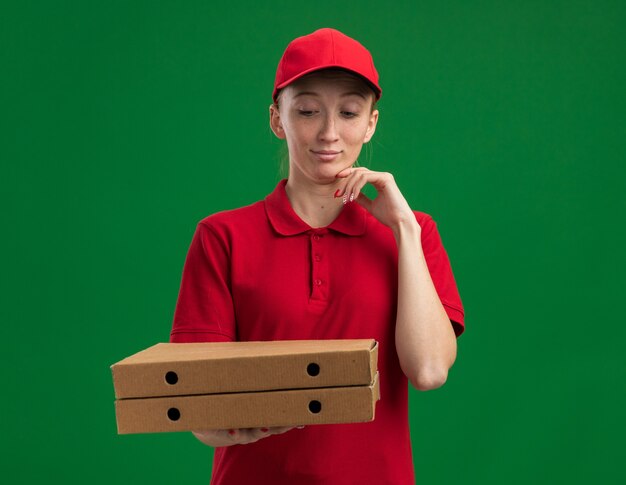 Young delivery girl in red uniform and cap holding pizza boxes looking at them with hand on chin thinking standing over green wall