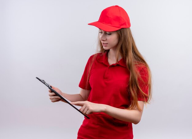 Young delivery girl in red uniform holding and putting finger on clipboard and looking at it on isolated white wall