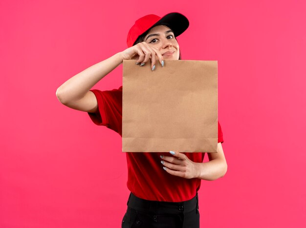 Young delivery girl wearing red polo shirt and cap holding paper package looking at camera with smile on face standing over pink background