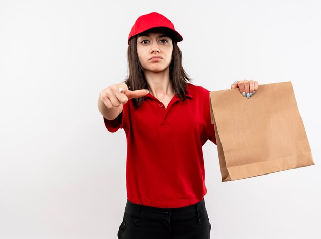 Young delivery girl wearing red uniform and cap holding paper package pointing with index finger at camera looking with serious face standing over white background