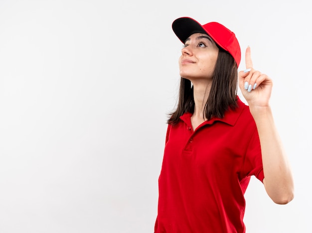 Free photo young delivery girl wearing red uniform and cap looking up showing index finger smiling confident standing over white background