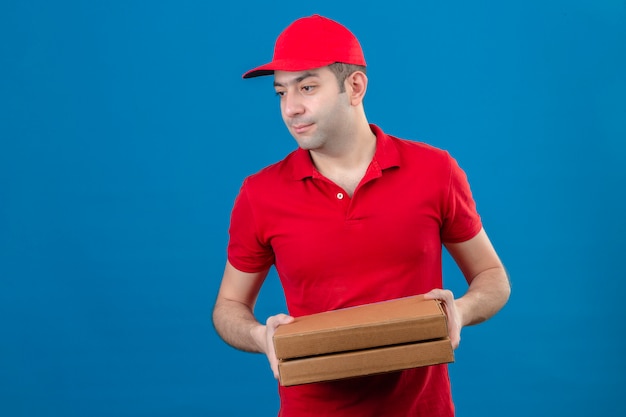 Young delivery man in red polo shirt and cap holding pizza boxes looking to the side with serious face over isolated blue wall