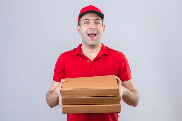 Young delivery man in red polo shirt and cap holding pizza boxes smiling cheerfully standing over isolated white wall