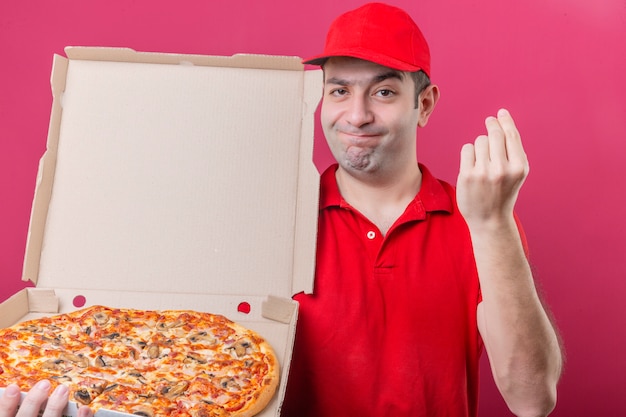 Free photo young delivery man in red polo shirt and cap standing with box of fresh pizza making delicious gesture with hand grins at camera over isolated pink background