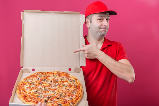 Free photo young delivery man in red polo shirt and cap standing with box of fresh pizza pointing to it with finger looking at camera convinced and confident over isolated pink background