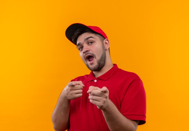 Young delivery man in red uniform and cap happy and positive pointing with fingers to camera