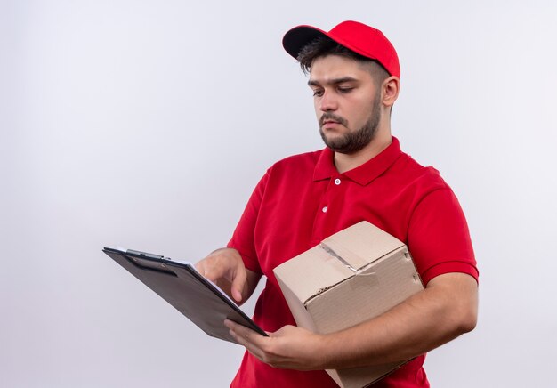 Young delivery man in red uniform and cap holding box package and clipboard with blank pages looking at them with serious face 