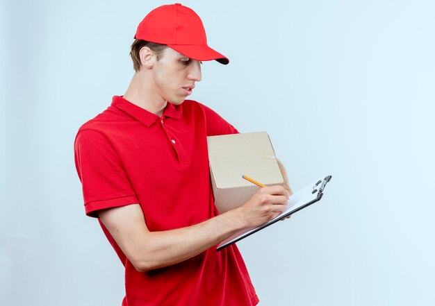 Young delivery man in red uniform and cap holding box package and clipboard writing something with serious face standing over white wall