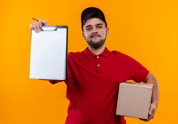 Young delivery man in red uniform and cap holding box package showing clipboard with blank pages asking for signature