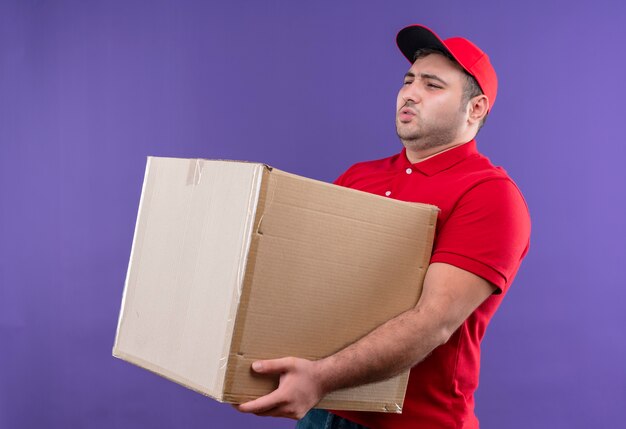 Young delivery man in red uniform and cap holding large cardboard box looking unwell suffering from heavy weight standing over purple wall