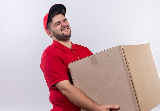 Young delivery man in red uniform and cap holding large cardboard box suffering from heavy weight 