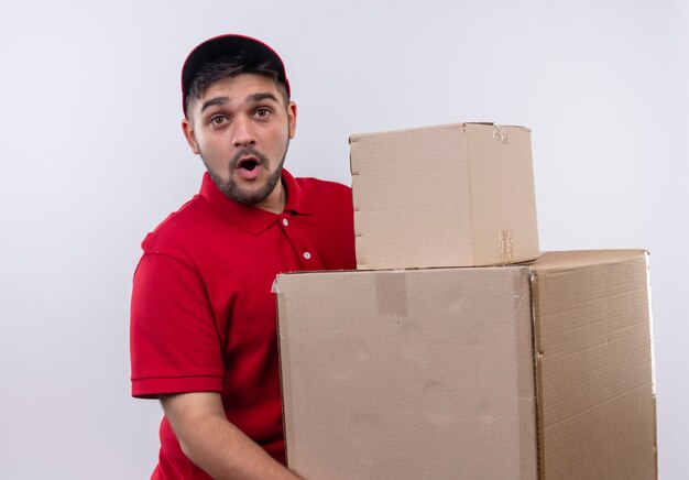 Young delivery man in red uniform and cap holding large cardboard boxes looking surprised and amazed 