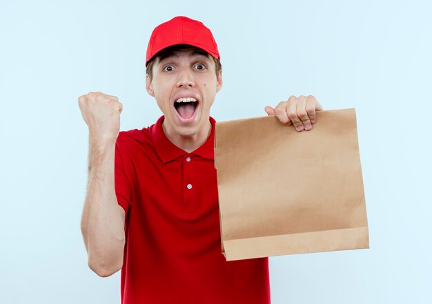 Young delivery man in red uniform and cap holding paper package clenching fist happy and excited standing over white wall