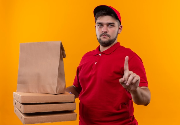 Young delivery man in red uniform and cap holding paper package and stack of pizza boxes showing index finger warning with serious face