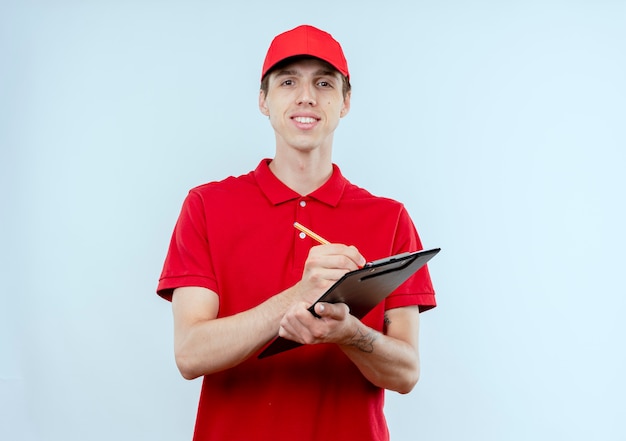 Young delivery man in red uniform and cap holding and pencile looking to the front with confident expression standing over white wall