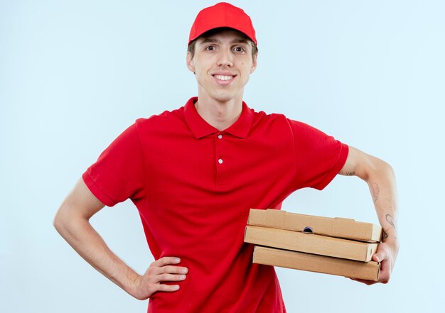 Young delivery man in red uniform and cap holding pizza boxes looking to the front with confident expression standing over white wall