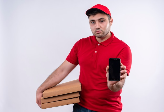 Young delivery man in red uniform and cap holding pizza boxes showing smartphone with confident expression standing over white wall