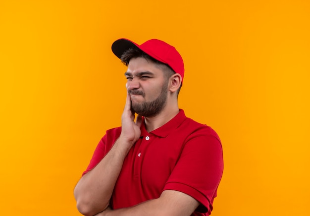 Young delivery man in red uniform and cap looking aside confused and very anxious