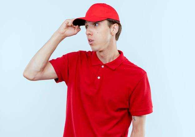 Young delivery man in red uniform and cap looking aside confused with hand oh his head standing over white wall