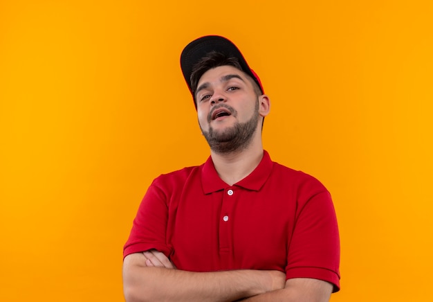 Young delivery man in red uniform and cap looking at camera with crossed arms on chest self-satisfied and proud