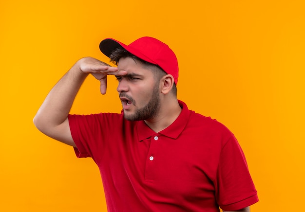 Free photo young delivery man in red uniform and cap looking far away with hand over head to look something