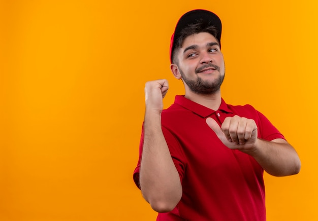 Young delivery man in red uniform and cap pointing back smiling confident