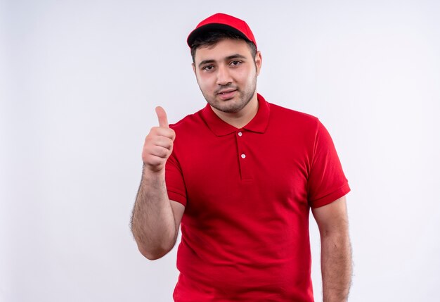 Young delivery man in red uniform and cap smiling cheerfully showing thumbs up standing over white wall