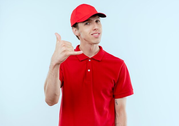 Young delivery man in red uniform and cap smiling confident making call me gesture standing over white wall