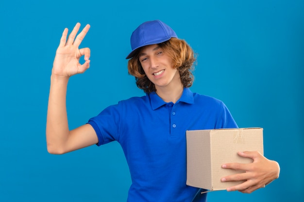 Young delivery man wearing blue polo shirt and cap holding cardboard box doing ok sign smiling friendly over isolated blue background