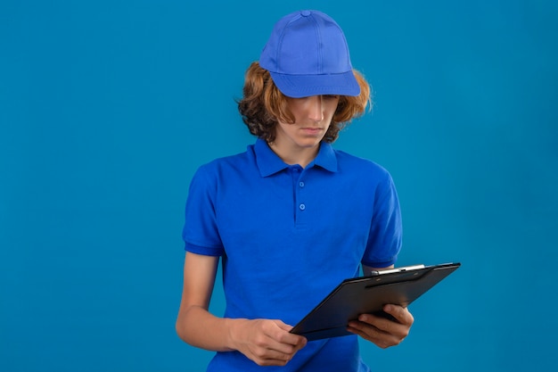 Young delivery man wearing blue polo shirt and cap looking at clipboard in his hand with unhappy face standing over isolated blue background