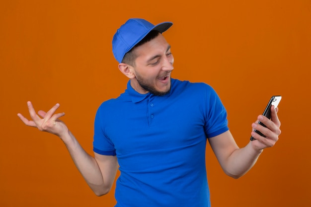 Young delivery man wearing blue polo shirt and cap looking at mobile phone in hand with happy face smiling cheerfully over isolated orange background