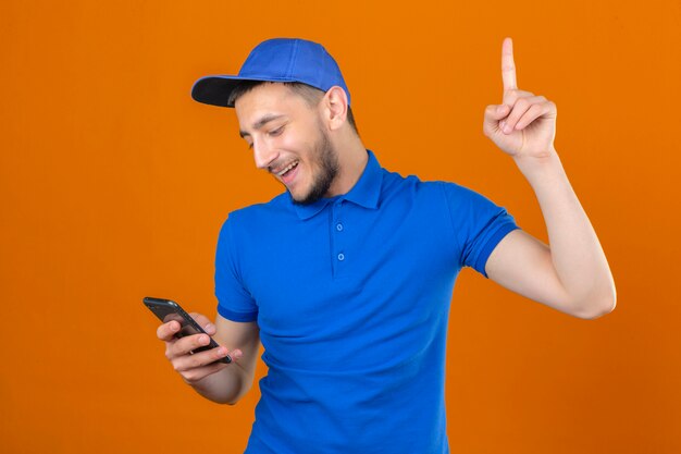 Young delivery man wearing blue polo shirt and cap standing with smartphone in hand pointing finger up smiling new idea concept over isolated orange background