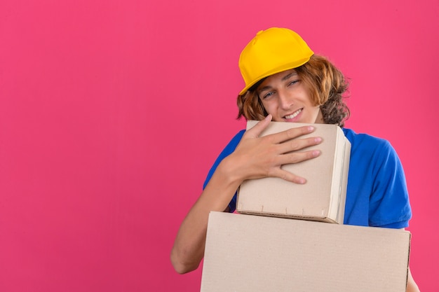 Free Photo young delivery man wearing blue polo shirt and yellow cap holding cardboard boxes dreaming embracing boxes smiling with happy face over isolated pink background