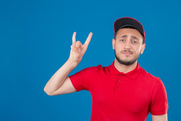 Young delivery man wearing red polo shirt and cap doing rock symbol looking confident over isolated blue background