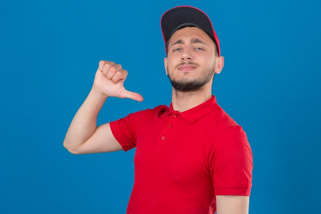 Young delivery man wearing red polo shirt and cap looking at camera smiling cheerfully proud and self-satisfied over isolated blue background