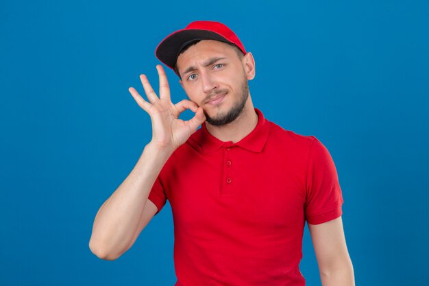 Young delivery man wearing red polo shirt and cap making silence gesture doing like closing his mouth with a zipper over isolated blue background