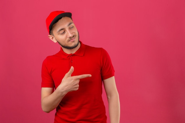 Free photo young delivery man wearing red polo shirt and cap pointing to the side to present something over isolated pink background