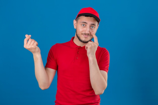 Young delivery man wearing red polo shirt and cap worried about money doing a money gesture with hand over isolated blue background