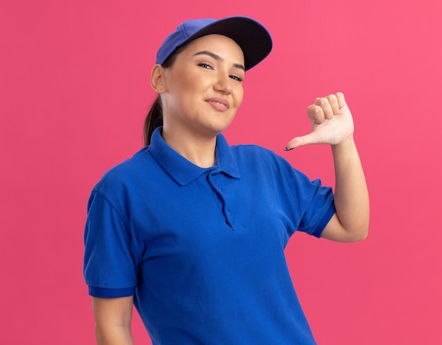 Young delivery woman in blue uniform and cap happy and confident pointing with thumb at herself standing over pink wall