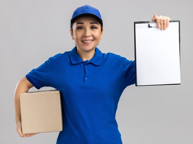 Young delivery woman in blue uniform and cap holding cardboard box showing clipboard with blank pages smiling confident  standing over white wall